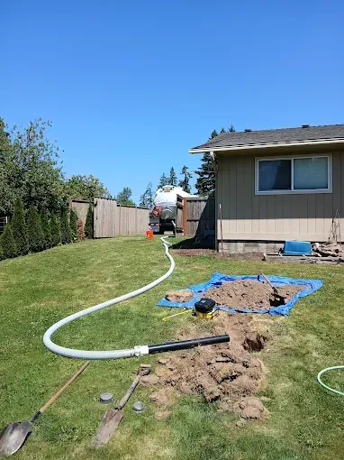 Backyard with a trench being dug, a septic tank in the distance, and a white hose. Sunny day.