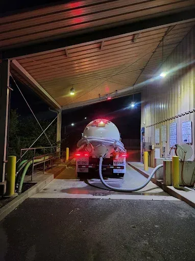Tanker truck under a canopy at night, connected to a hose. Red taillights are on.