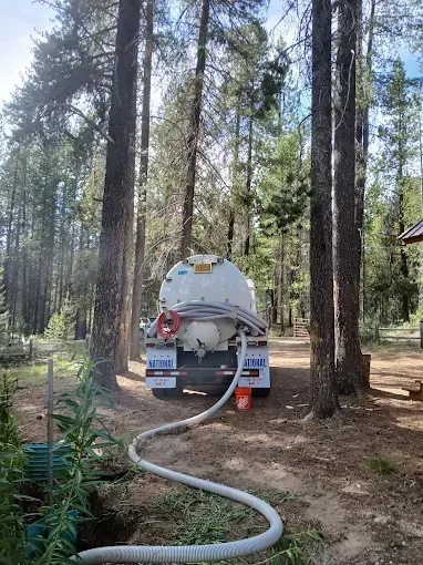 A water tanker truck parked in a forest, discharging water through a hose into a dark opening in the ground.