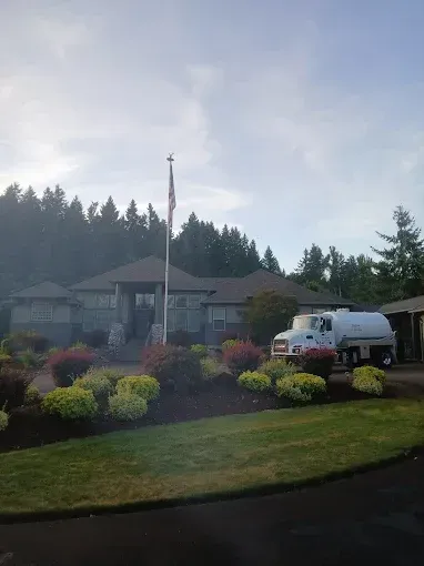 A light-colored building with a flagpole, a water truck, and green landscaping under a cloudy sky.