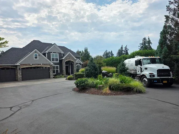White tanker truck parked in front of a large gray house with black garage doors and a circular driveway.