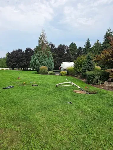 Lush green lawn with trees, bushes, and a tanker truck in the background. A white hose lies across the grass.