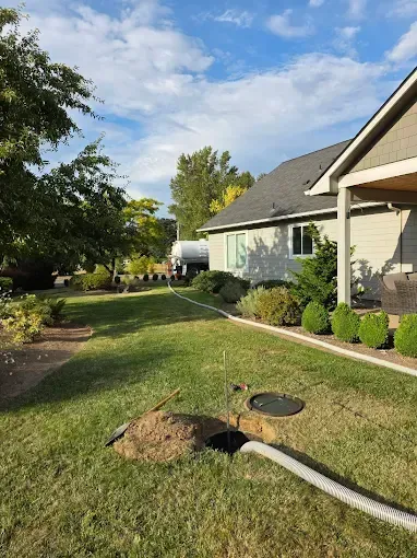 A septic tank being serviced with a hose laid across a grassy yard next to a house under a cloudy sky.