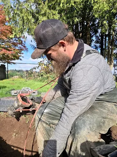 Man with a beard in a gray sweatshirt uses a grinder near an open trench outdoors on a sunny day.