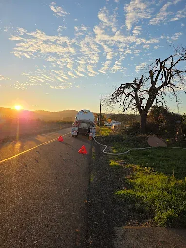 Tanker truck parked on roadside, orange cones, hose extending, sunrise, tree.