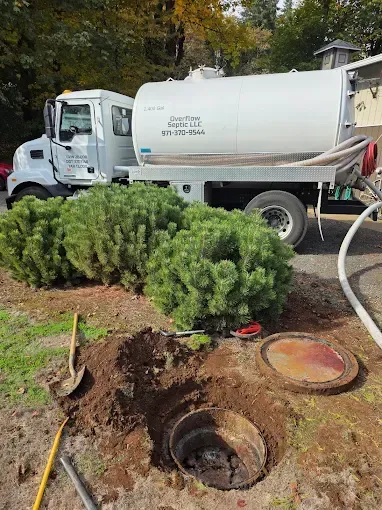 Septic tank being pumped by a white truck on a grassy area, bushes nearby, and an open tank.