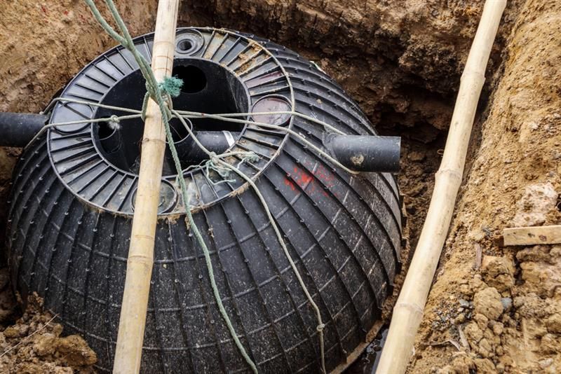 Black, ribbed septic tank partially buried in a brown earth trench with connecting pipes.
