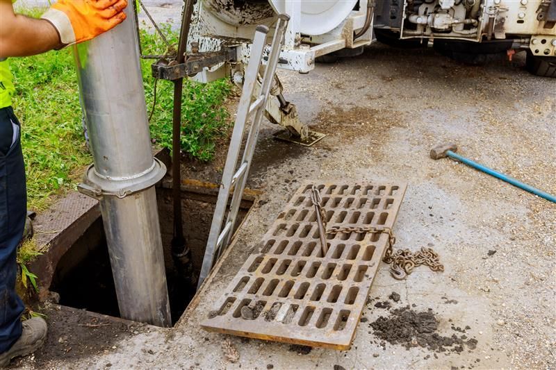 Man working on a sewer; orange gloves, metal pipe, open manhole, ladder, grate.