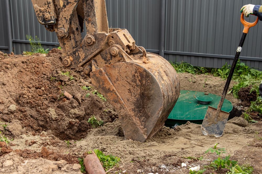 An excavator bucket digs near a septic tank, and a shovel sits nearby.
