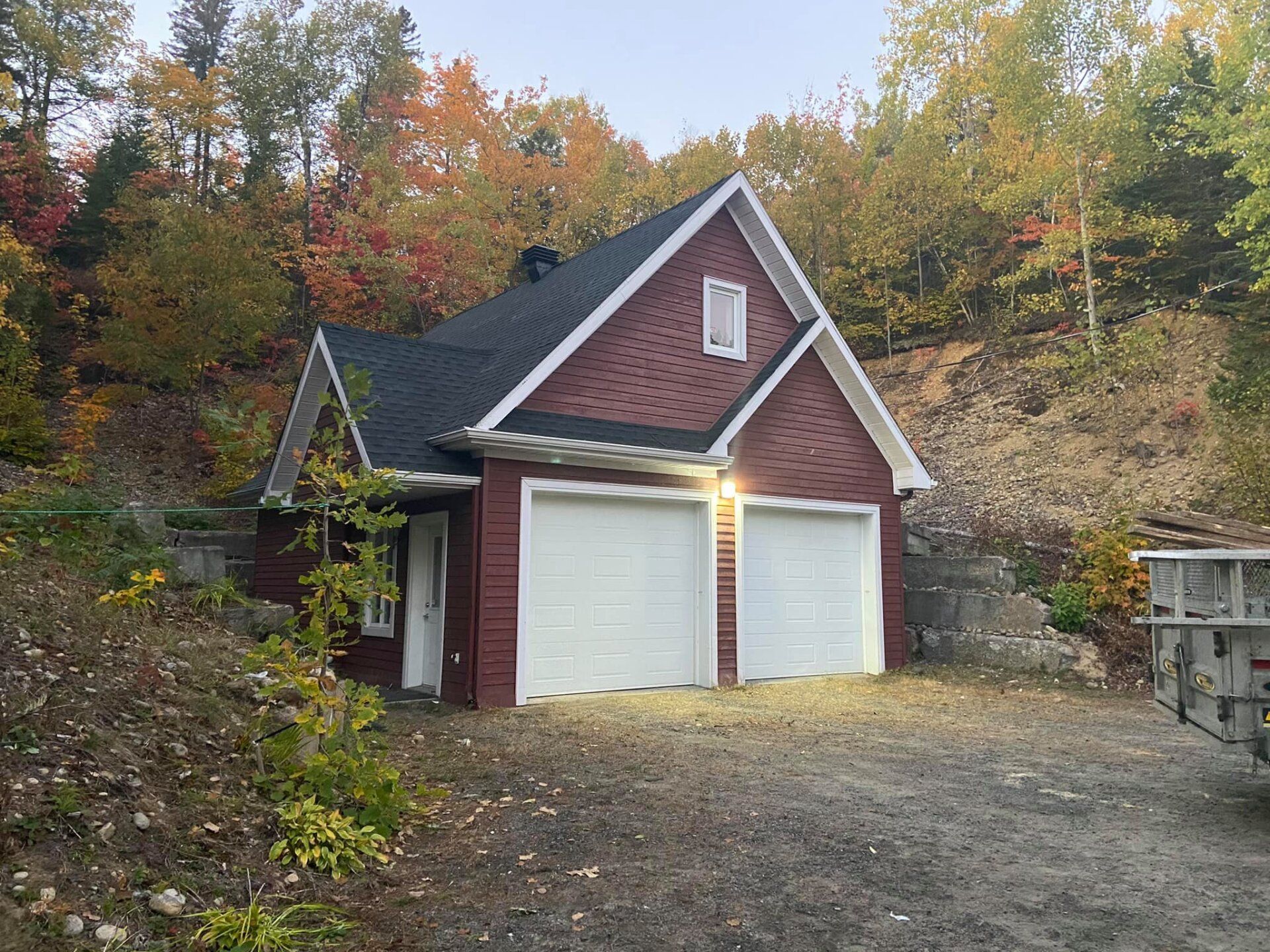Un garage en brique rouge avec deux portes de garage blanches est entouré d'arbres.