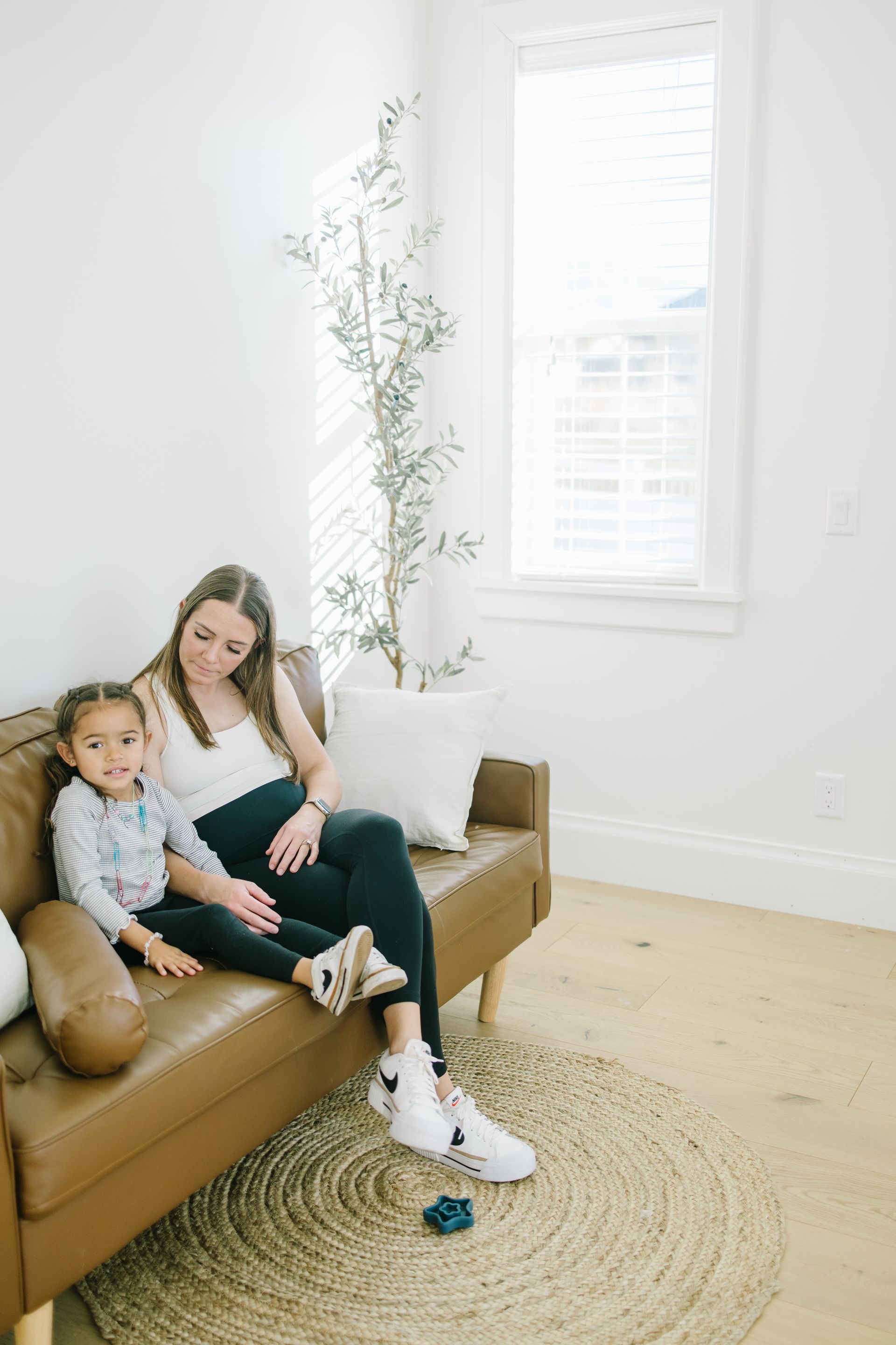 Woman and child sitting on a brown sofa in a bright living room with a woven rug and large window