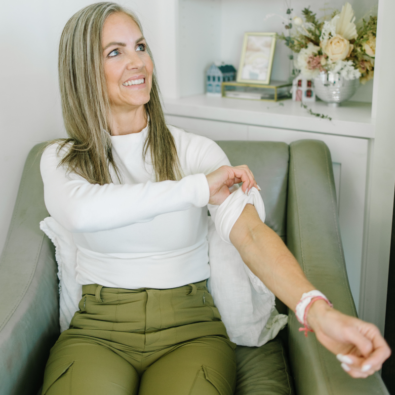 Woman sitting on a chair, rolling up her sleeve to reveal a bandage on her upper arm.