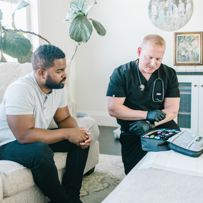 A nurse prepares a medical kit while speaking with a seated patient in a bright living room.