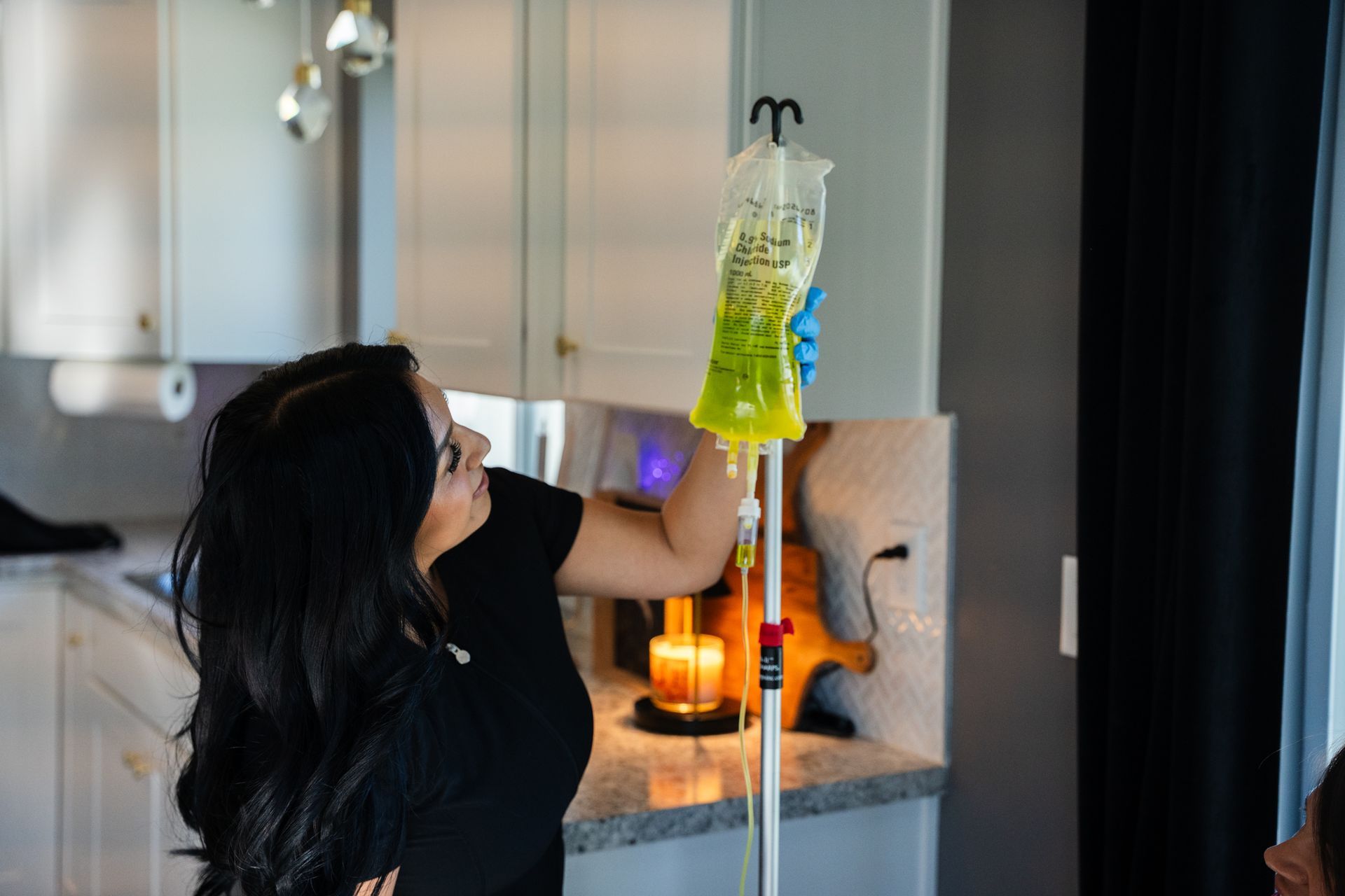 Person holding up a yellow IV bag in a kitchen near a lit candle