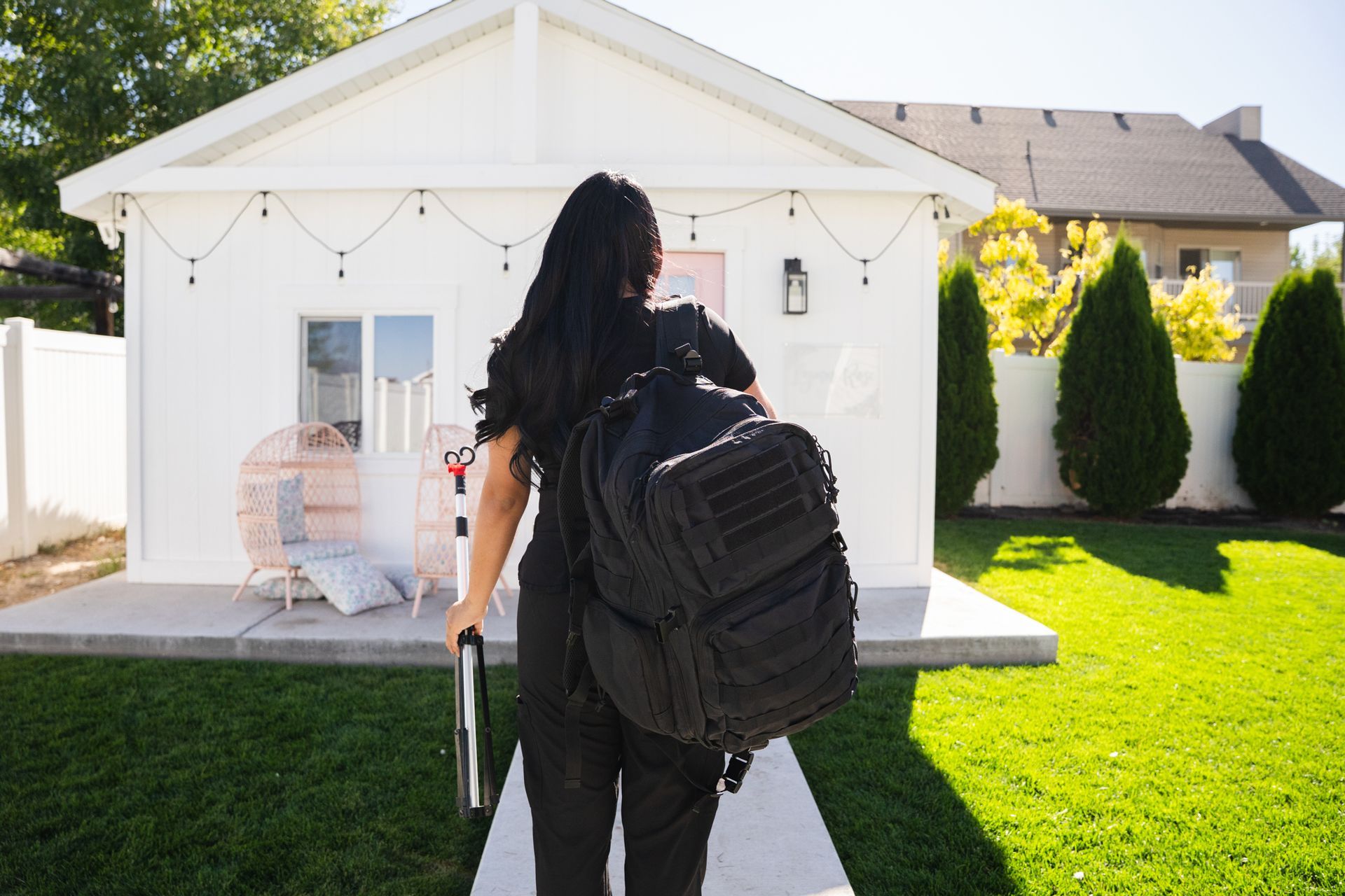 Person walking toward a small white house carrying a backpack and suitcase in a sunny yard.