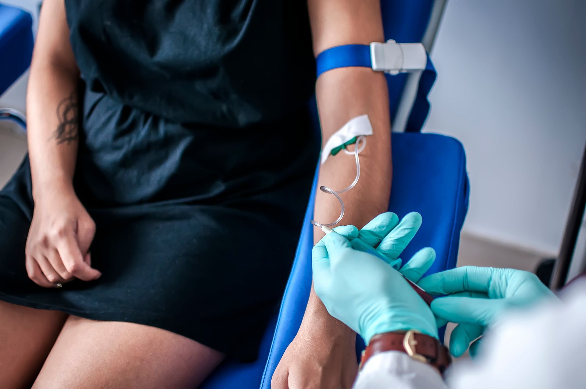 A healthcare worker in blue gloves performs a venipuncture on a person's arm using a needle and a blue tourniquet.