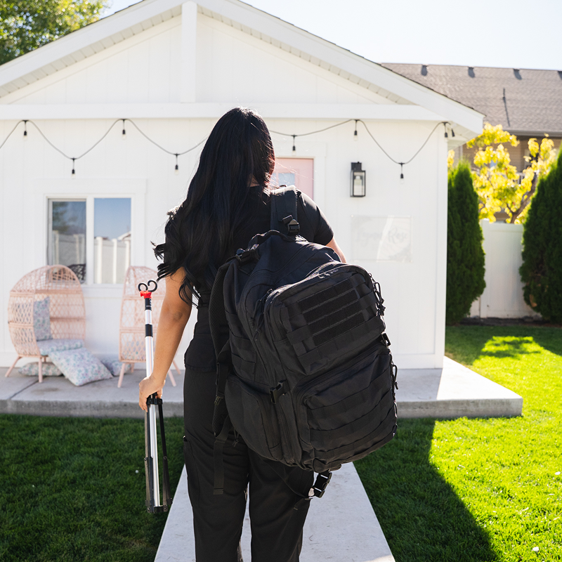 Person carrying a large black backpack and rolling luggage toward a small white house with string lights.