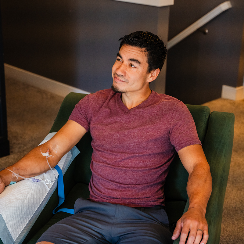 Man receiving IV treatment, sitting in a green chair, wearing maroon shirt and gray shorts, looking away.