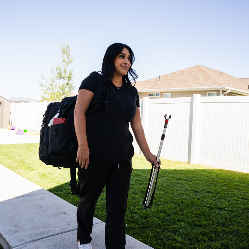 Person walking on a sidewalk with a black backpack, holding a tripod in a sunny suburban yard.