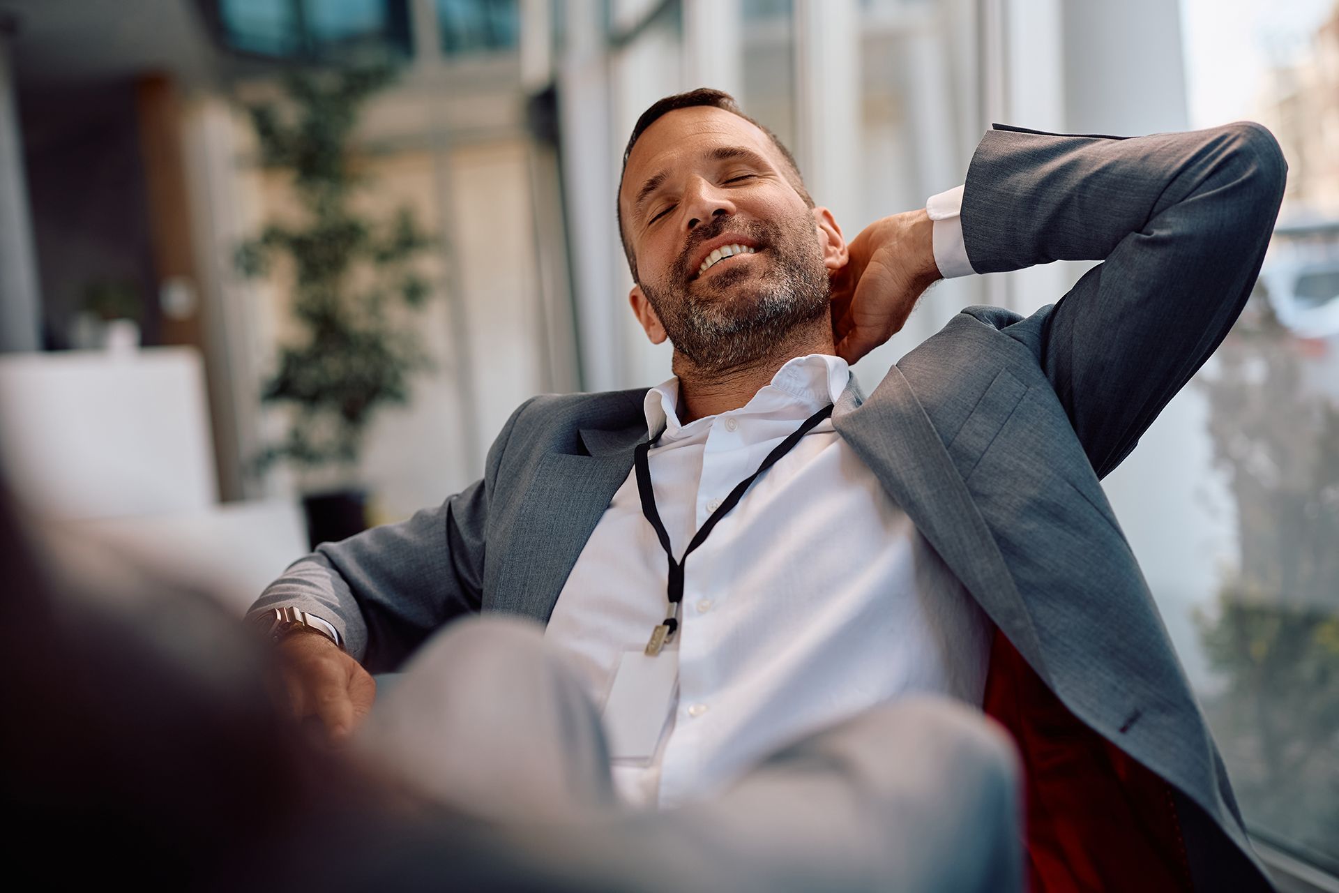 Relaxed man in a blazer reclining in an office, smiling with hands behind his head