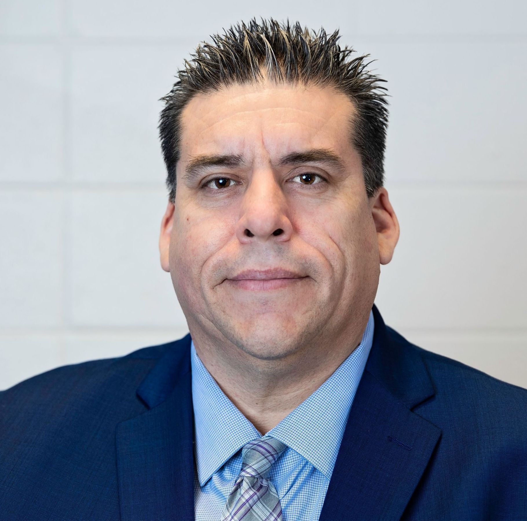 Man in a blue suit and patterned tie, with short dark hair, looking forward. White background.