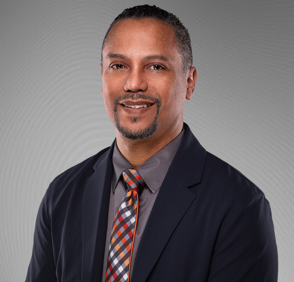 Man in blazer and tie, smiling against a grey background.