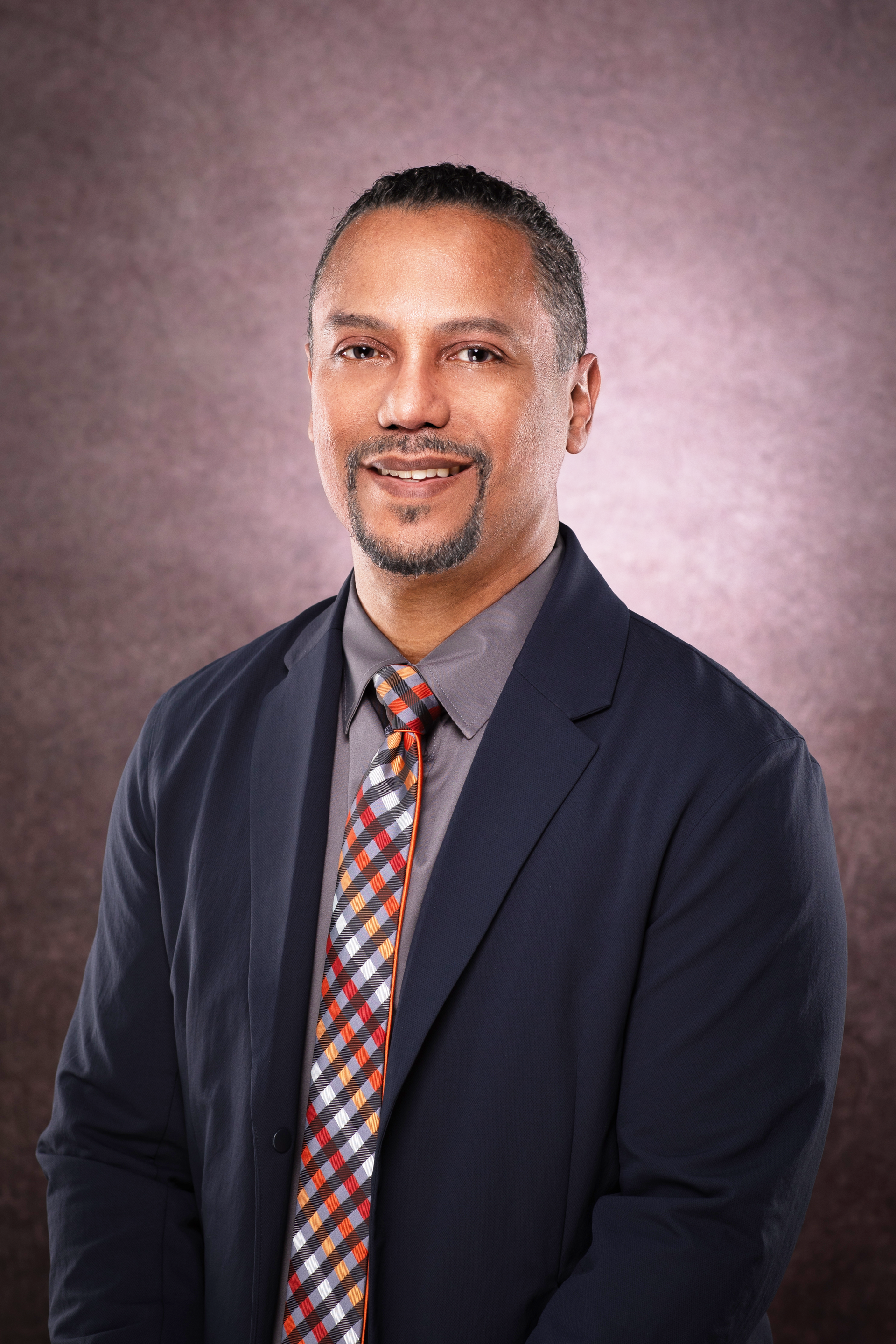 Man in a suit smiles, looking at the camera. He has a colorful tie and a brown background.
