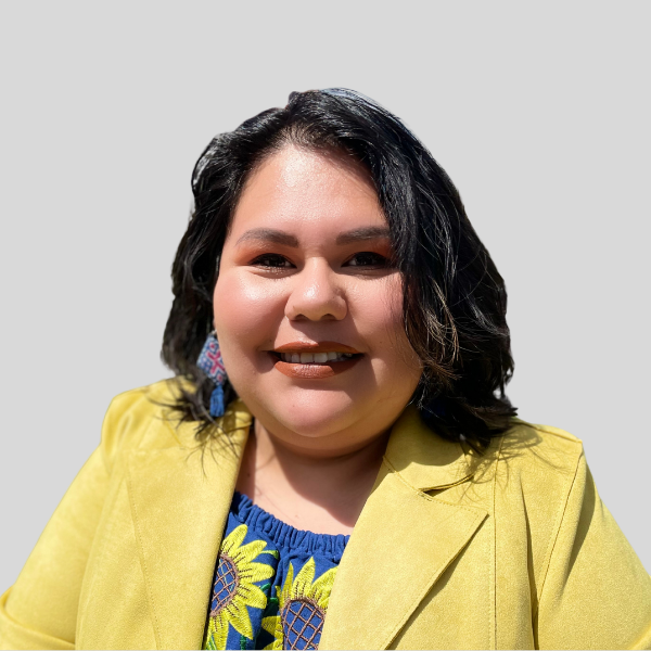 Woman with dark hair and smile wearing a yellow blazer and sunflower top; on a light gray background.