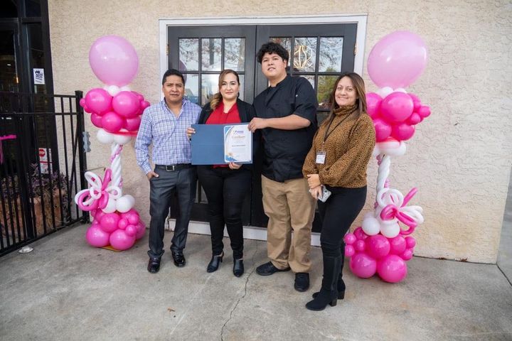 Four people stand outside a building, holding a certificate. Pink and white balloon decorations flank the doorway.