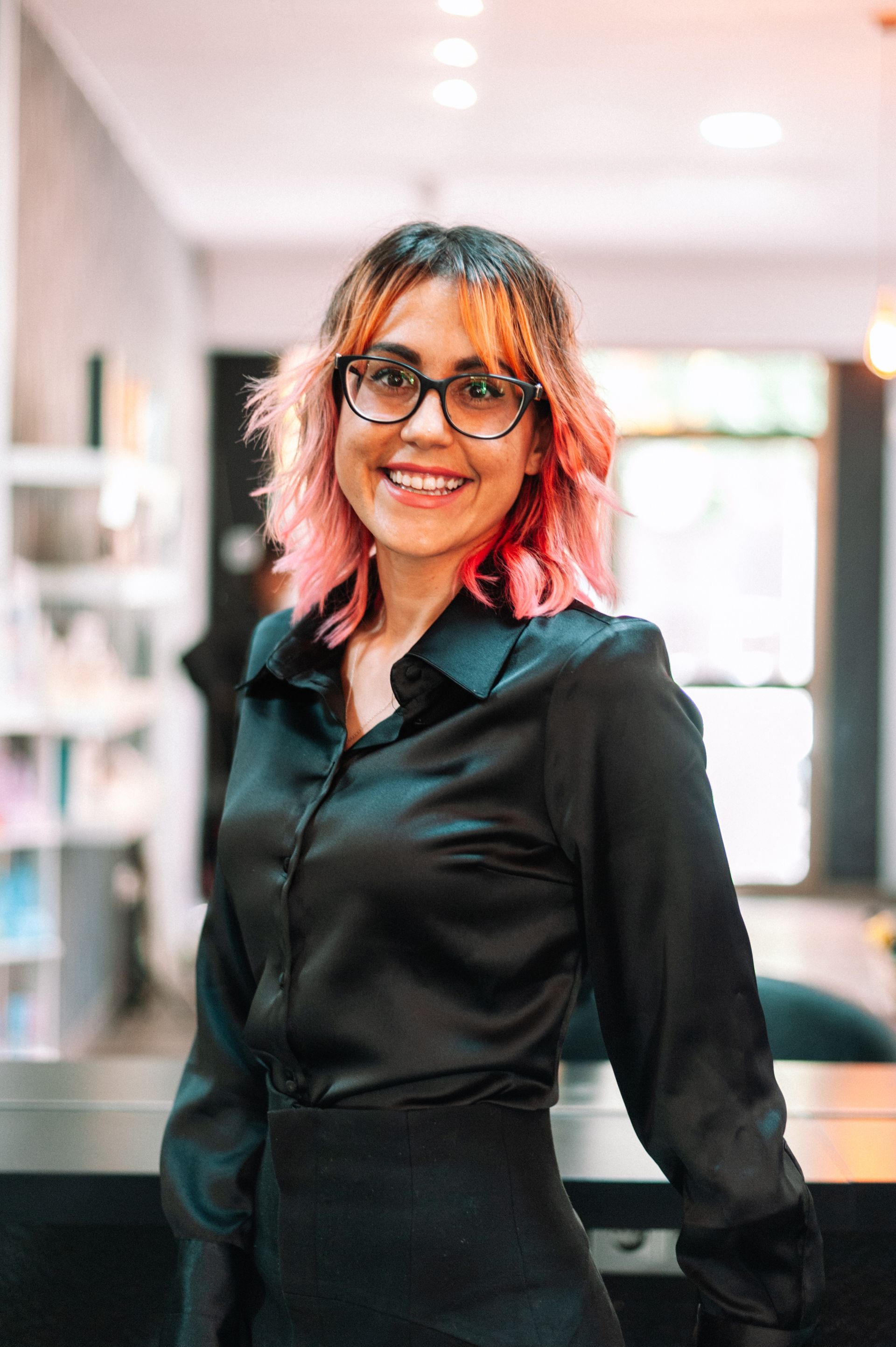 Woman with pink hair and glasses smiles in a salon, wearing a black shirt.