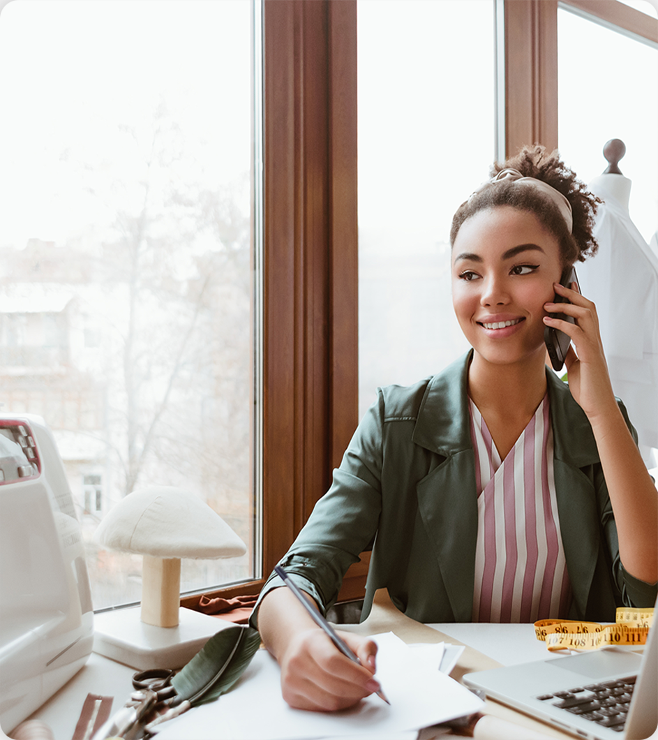 Woman with curly hair smiles while on a call, writing at a desk near a window.