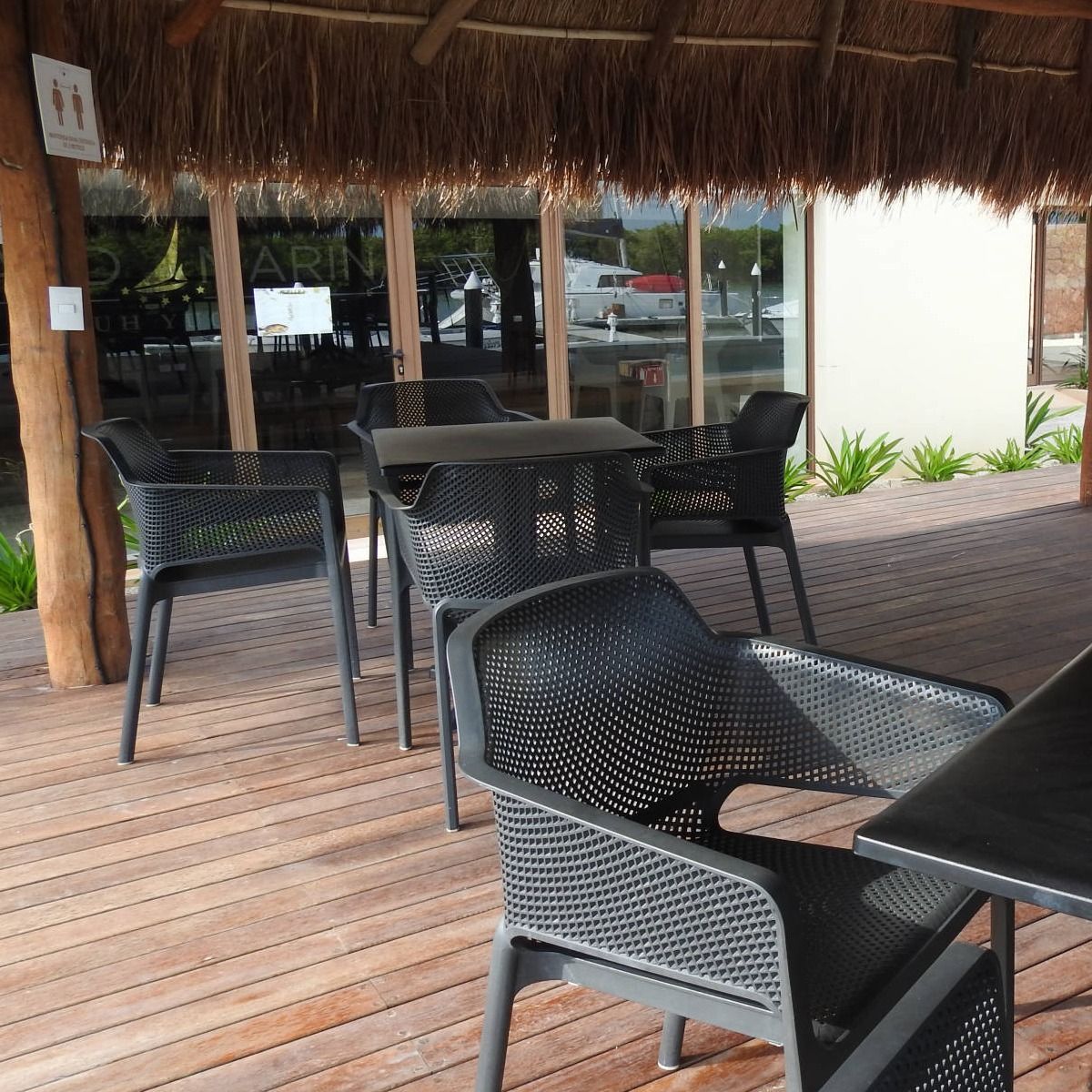 Una terraza de madera con sillas y mesas bajo un techo de paja.