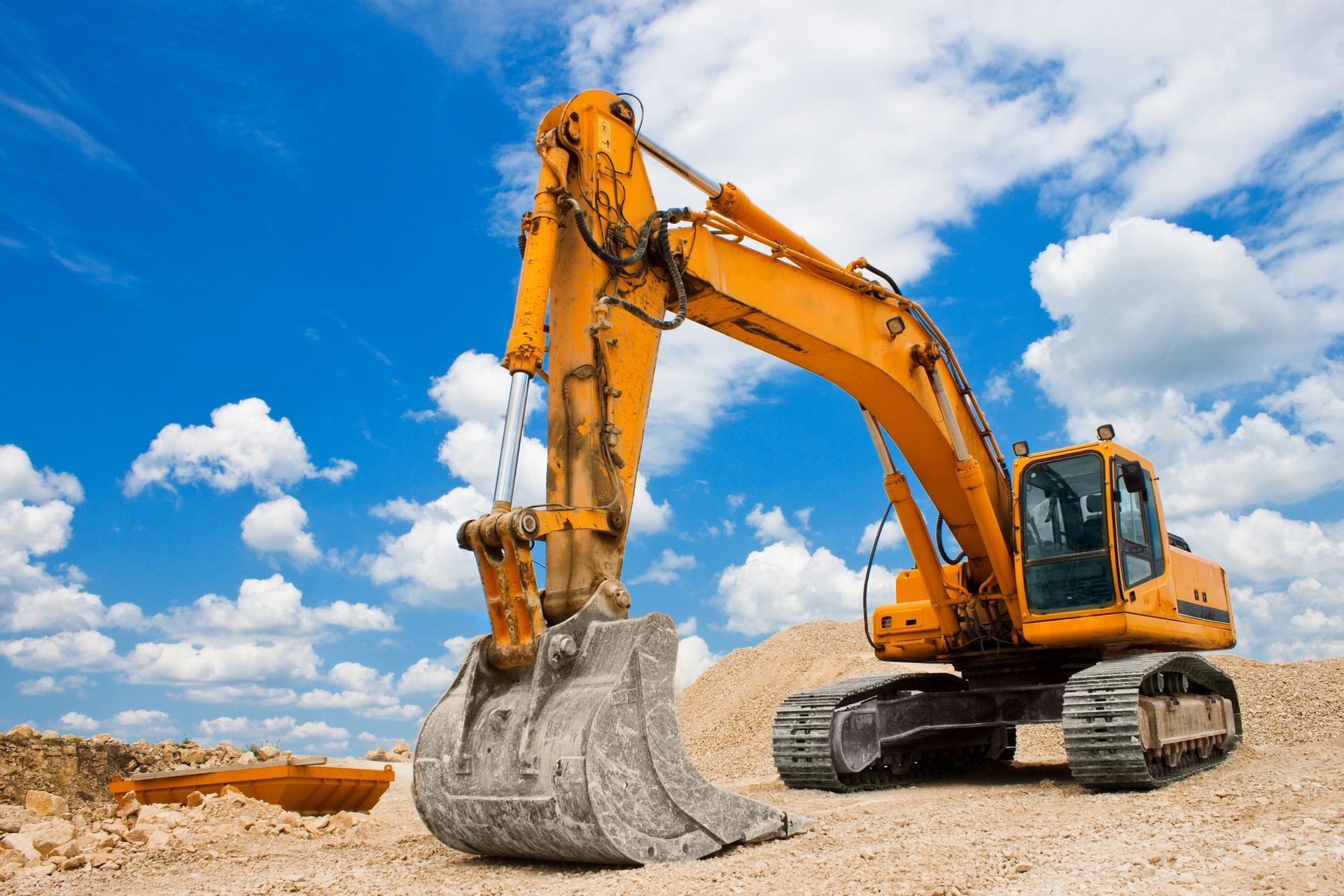 Excavadora amarilla en un terreno baldío bajo un cielo azul con nubes.
