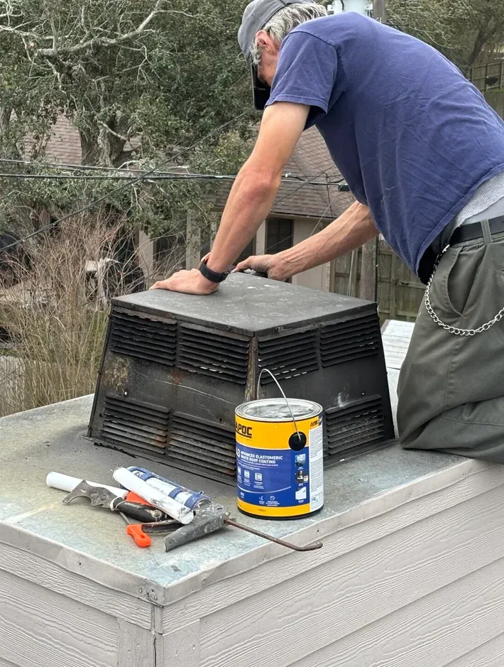 Man on a rooftop, working on a metal vent. Supplies, including paint can, are nearby.