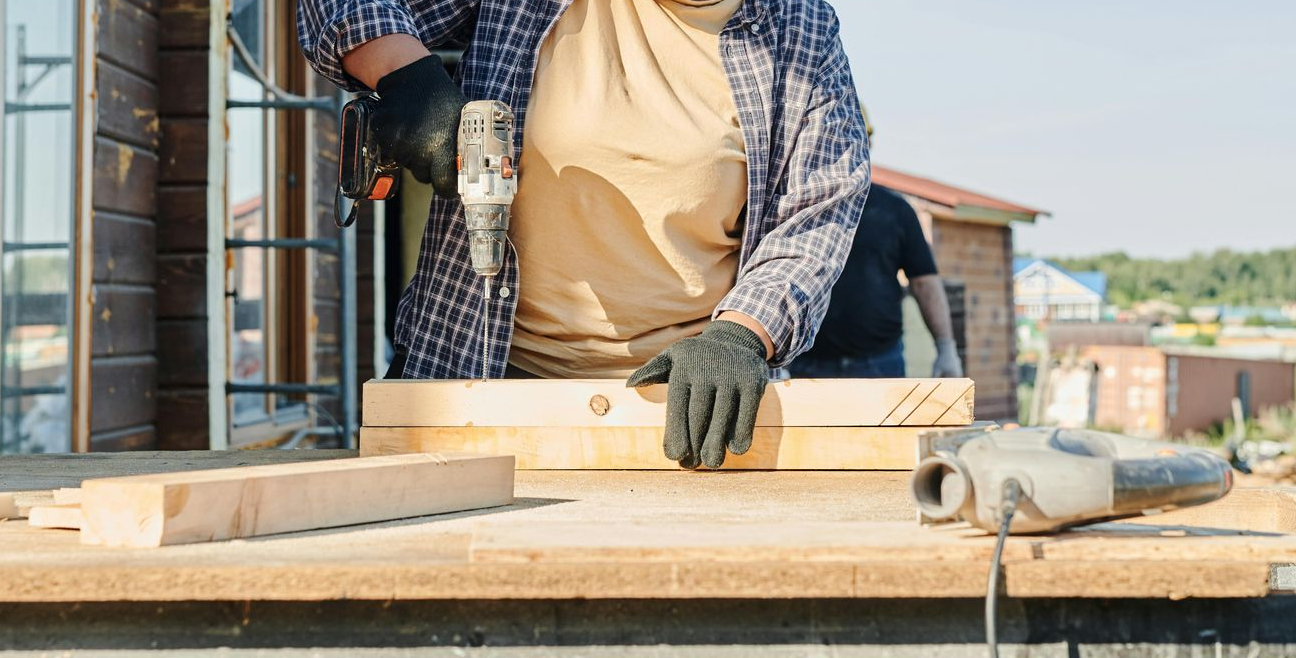 Person using a drill to work on wooden boards outdoors on a sunny day.