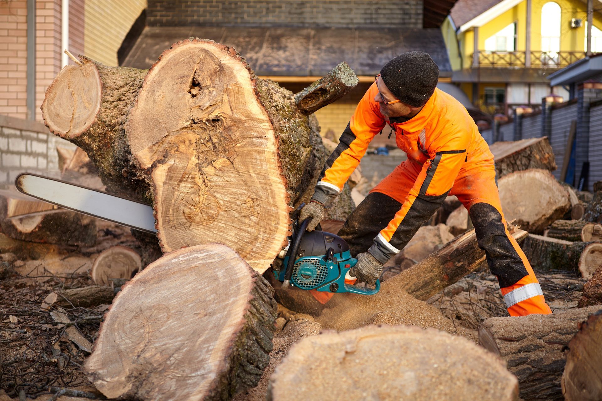 Worker in orange safety gear cutting large logs with a chainsaw in a wood yard