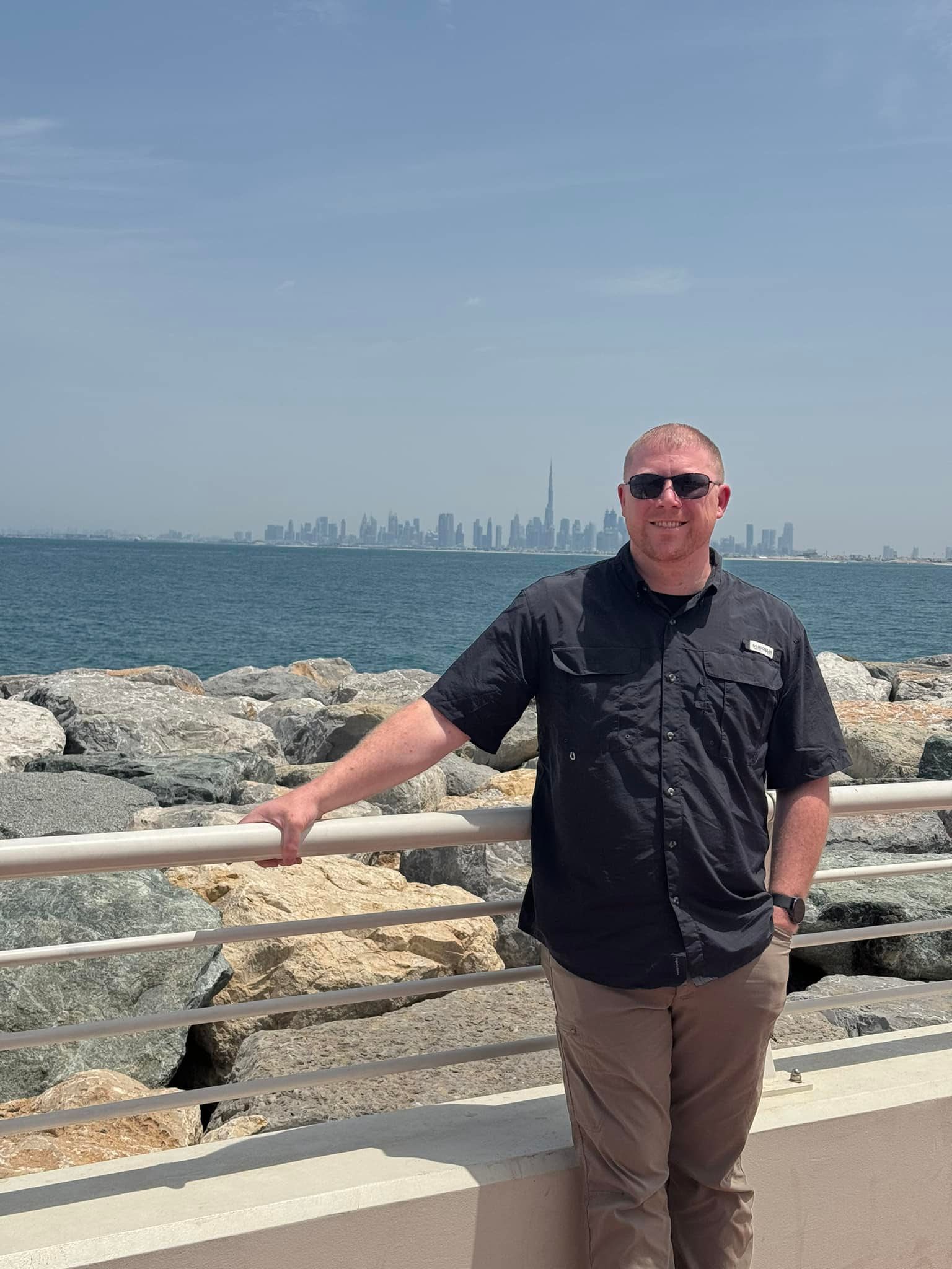 gentleman standing in front of rocks lining the ocean ; with a city skyline in the distance; leaning on a white fence