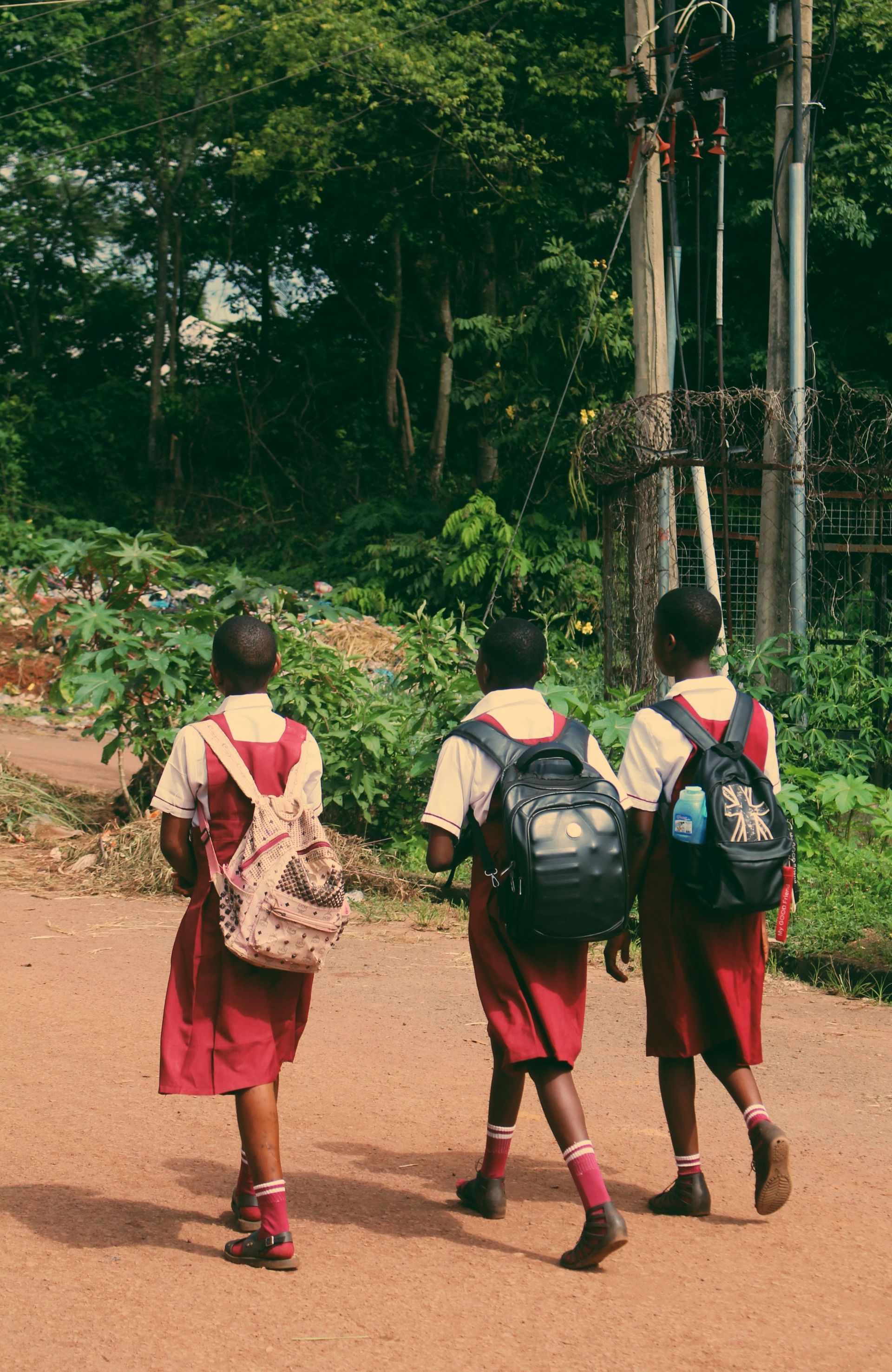 Three students in red uniforms walk along a dirt path with backpacks in a forested area.