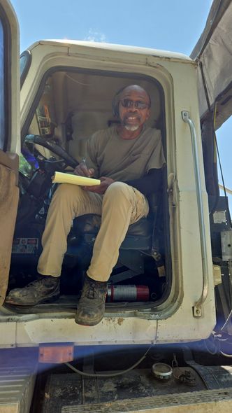 Man sitting inside an open white work van doorway, holding a tool in bright daylight.