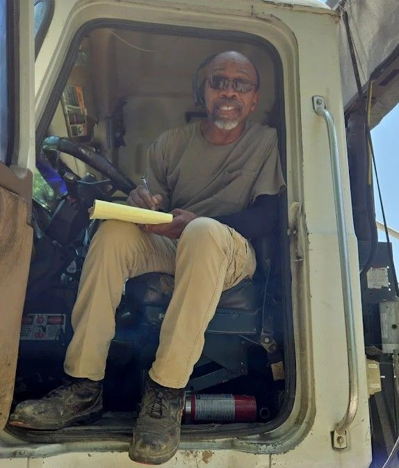 Man sitting in the cab doorway of a truck, holding a clipboard and smiling.