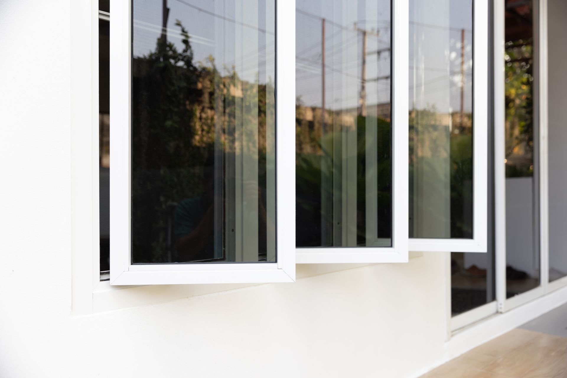 White-framed windows open in a white building, reflecting trees and sky.