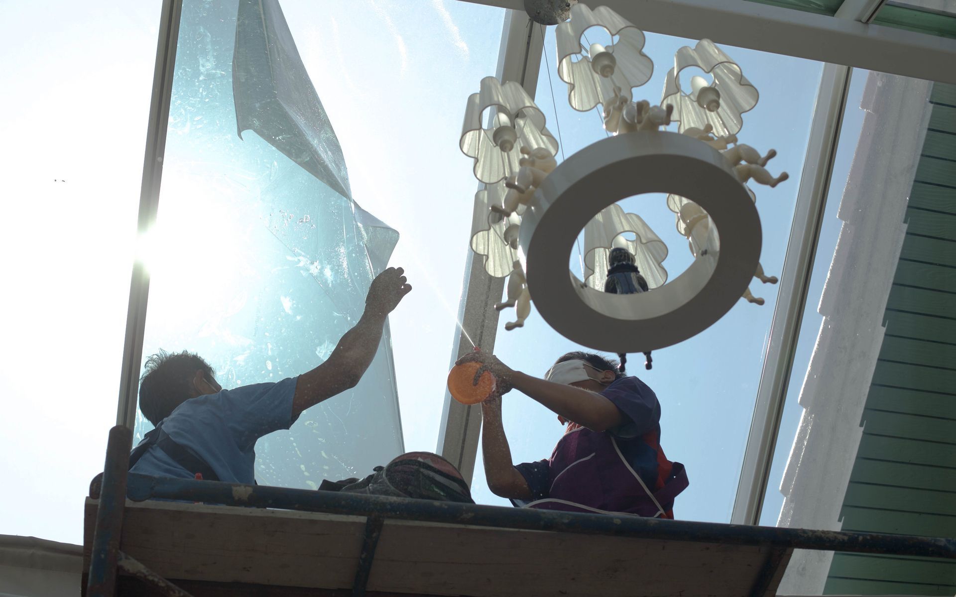 Two people install window film on a skylight, working from a platform. Bright sunlight.