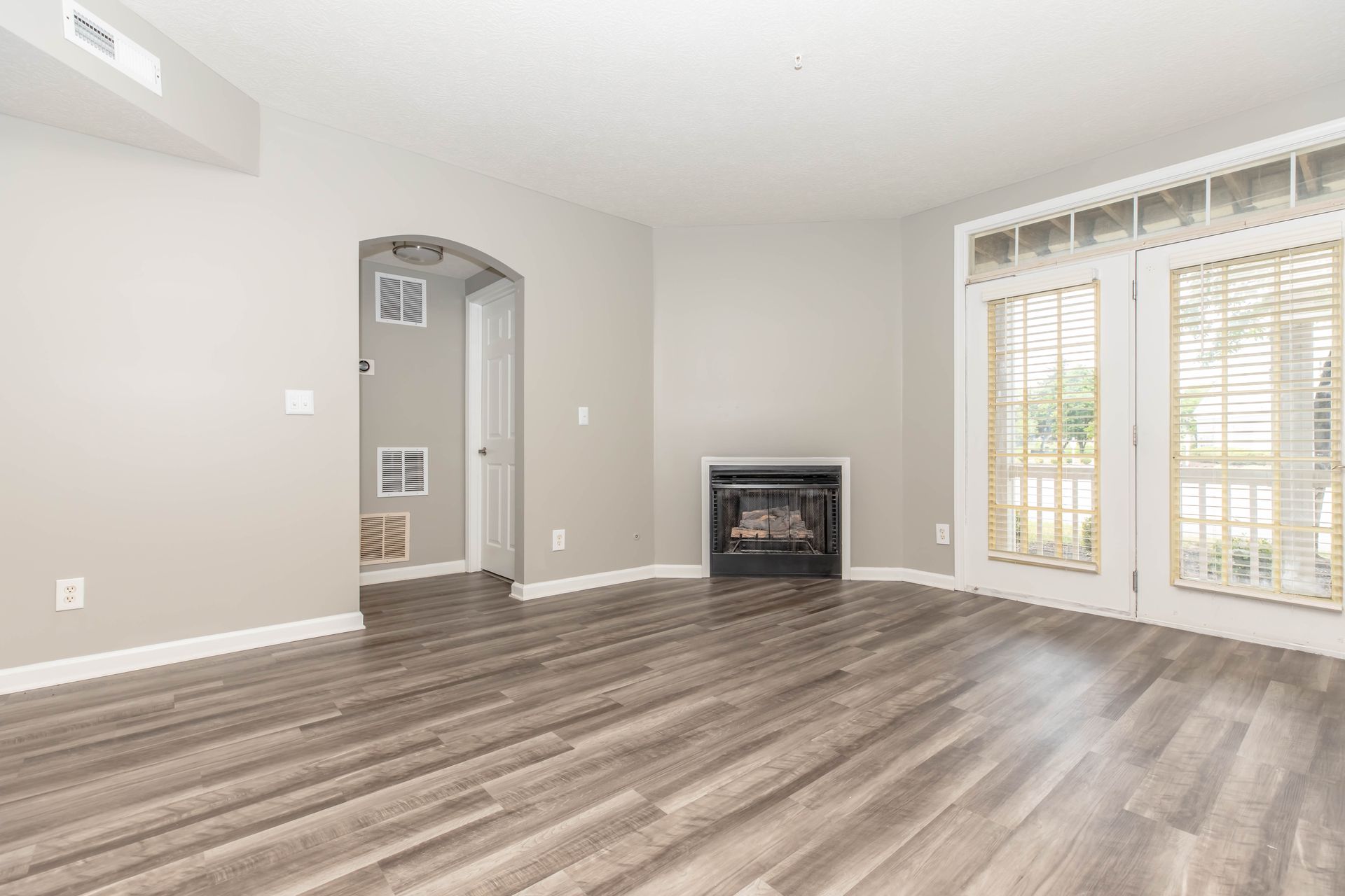 Empty living room with gray wood floors, white walls, a fireplace, and French doors with shutters at Quarry Apartments for Rent in Columbus, OH.