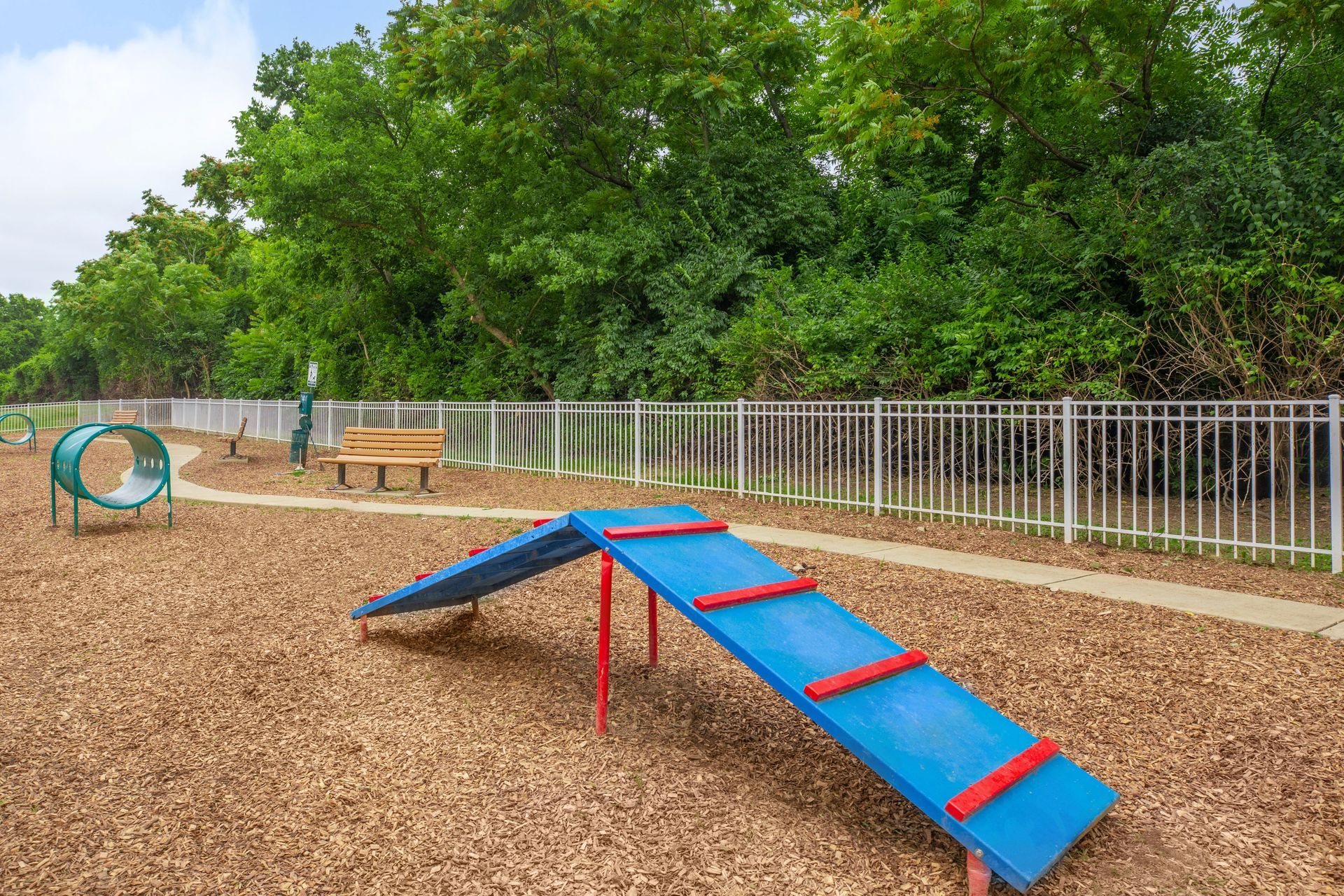 Outdoor dog agility course with a blue-and-red seesaw ramp, tunnel, and bench on wood chips under trees at Quarry Apartments for Rent in Columbus, OH.
