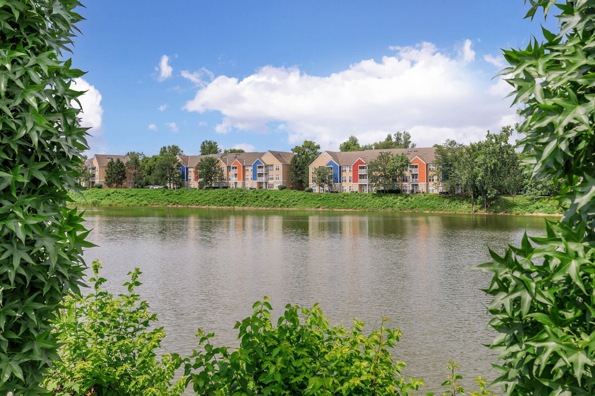 Lake view framed by green foliage, with apartment buildings across calm water under a blue sky at Quarry Apartments for Rent in Columbus, OH.