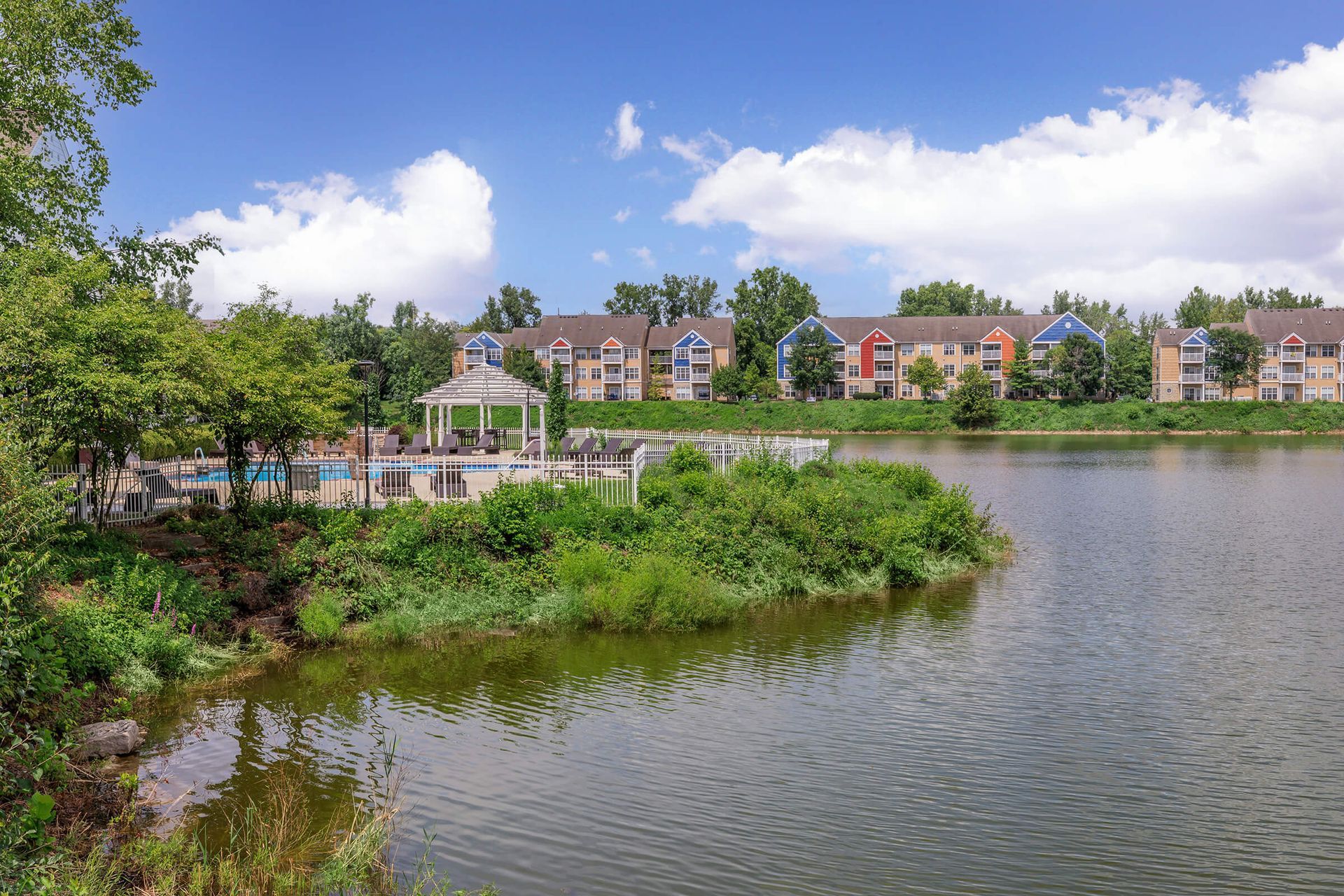 Calm lakeside view with trees, grassy shore, and apartment buildings under a bright blue sky at Quarry Apartments for Rent in Columbus, OH.