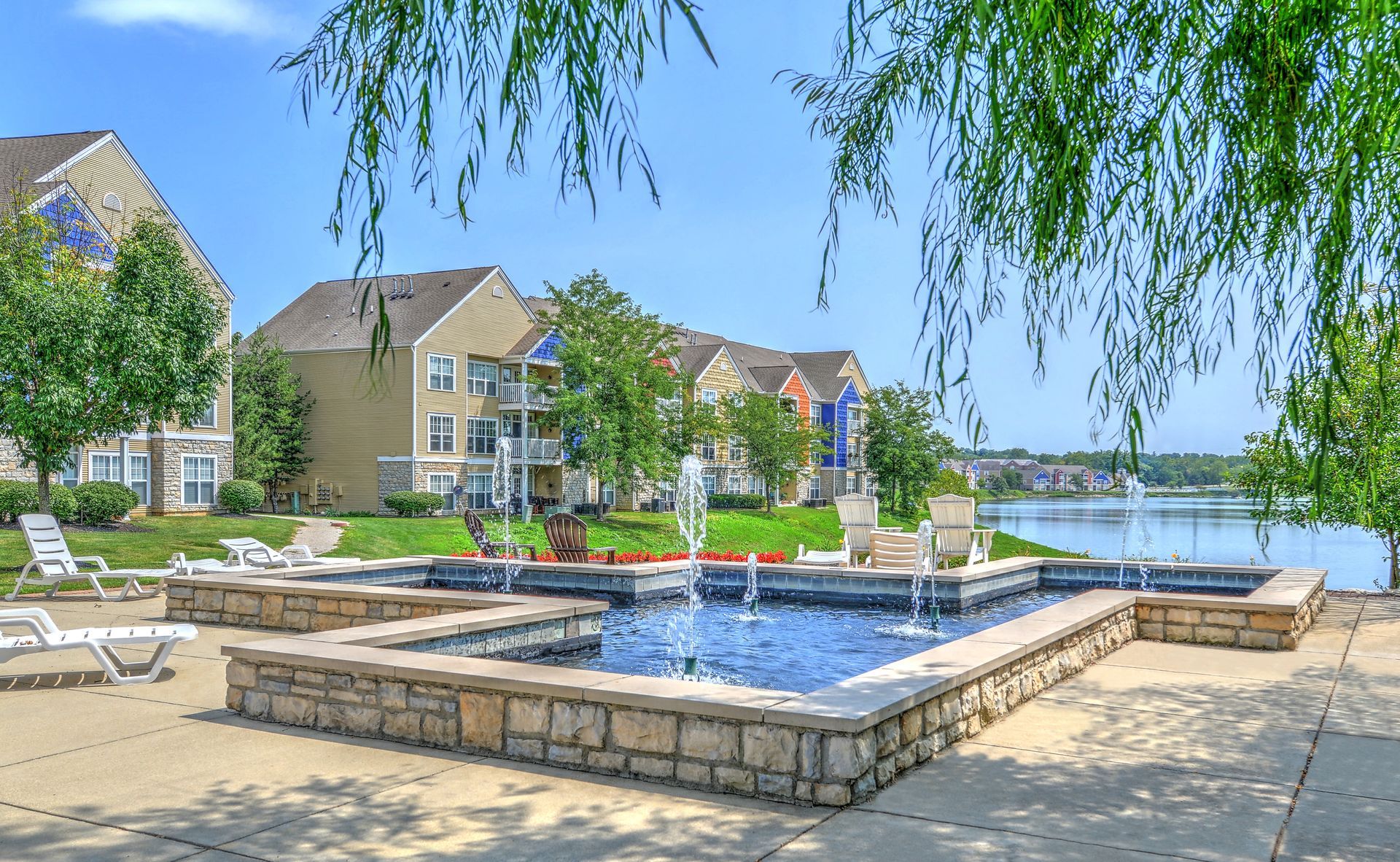 Courtyard with blue fountain, patio seating, lakeside apartments, and willow branches overhead at Quarry Apartments for Rent in Columbus, OH.