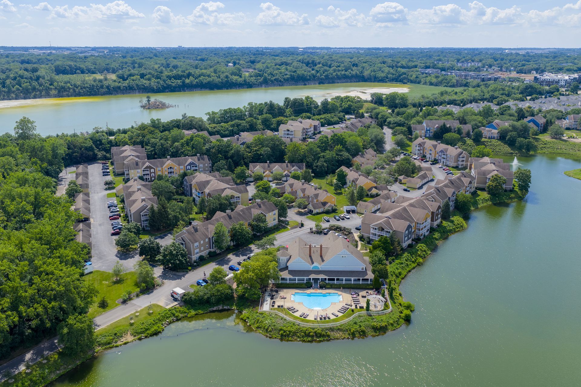 Aerial view of a waterfront apartment complex with a pool, lake, and surrounding trees at Quarry Apartments for Rent in Columbus, OH.