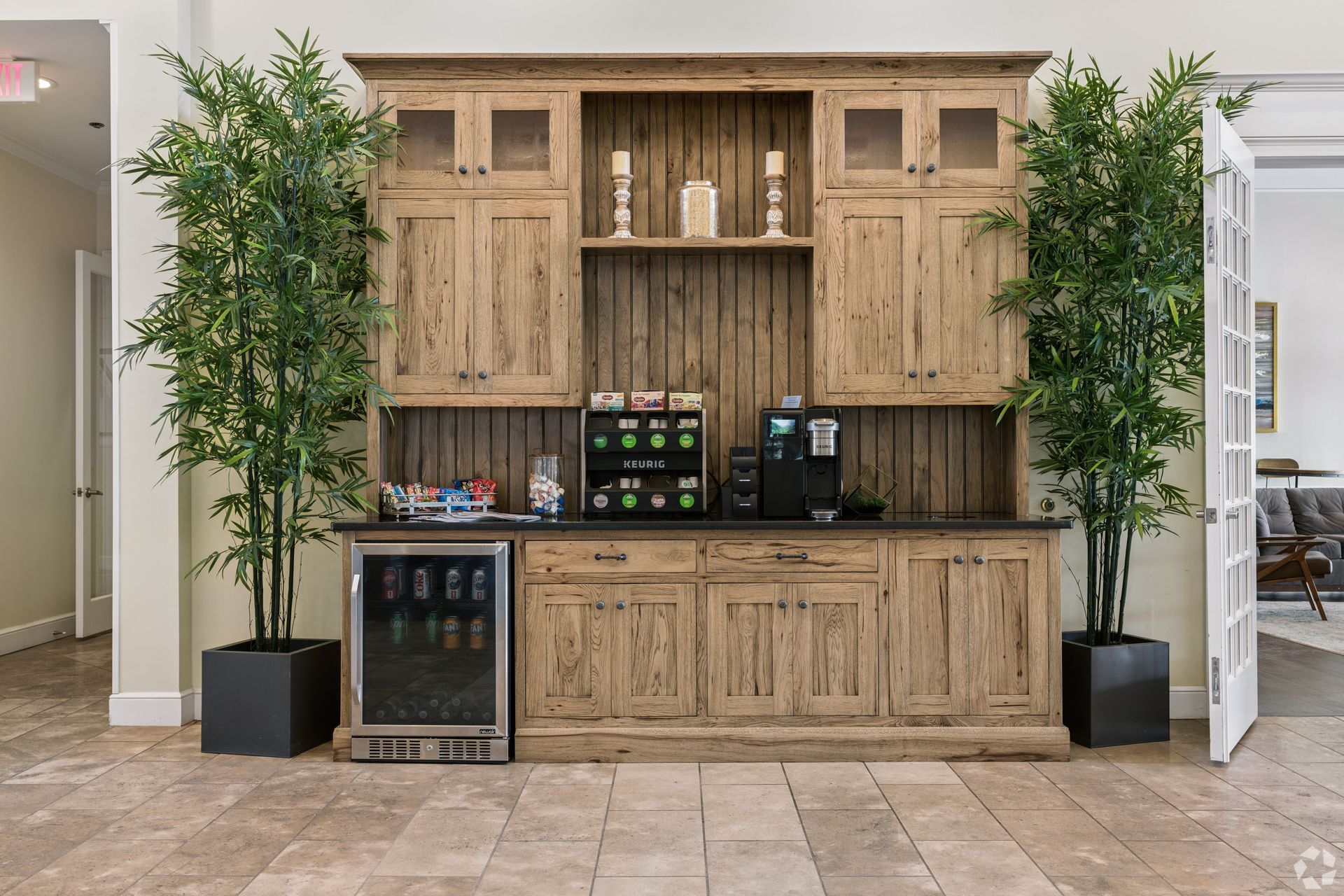 Wood-paneled beverage station with two potted trees, drink dispensers, and a small fridge in a tiled lobby at Quarry Apartments for Rent in Columbus, OH.