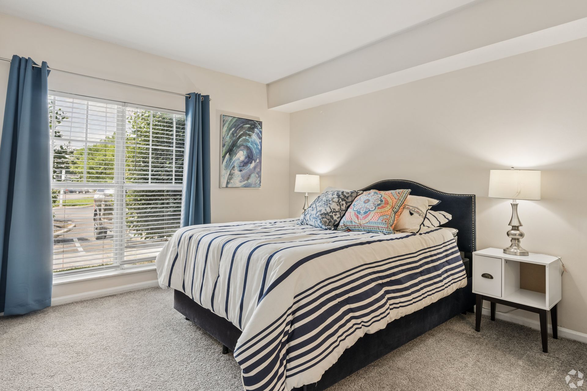 Bright bedroom with a striped bedspread, two nightstands, lamps, and a large window with blue curtains at Quarry Apartments for Rent in Columbus, OH.